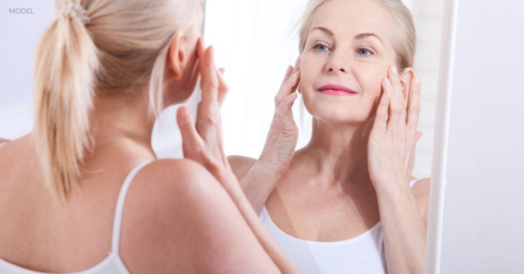 Woman with clear, youthful skin (model) looking in the mirror with hands held up to either side of her face.