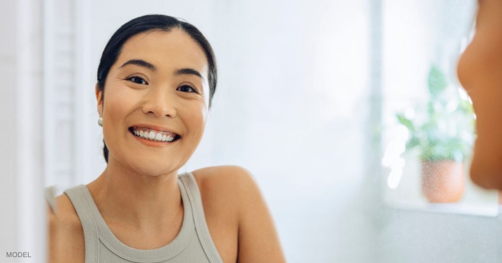 Woman with glowing beautiful skin (model) smiles while looking in the bathroom mirror