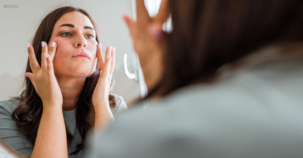 Woman exhibiting some redness of the face (model) examining her face closely in the mirror.