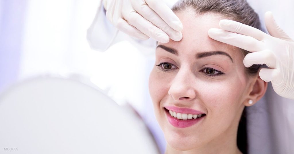 A young woman looks at herself in the mirror as a doctor examines her facial features (MODELS)